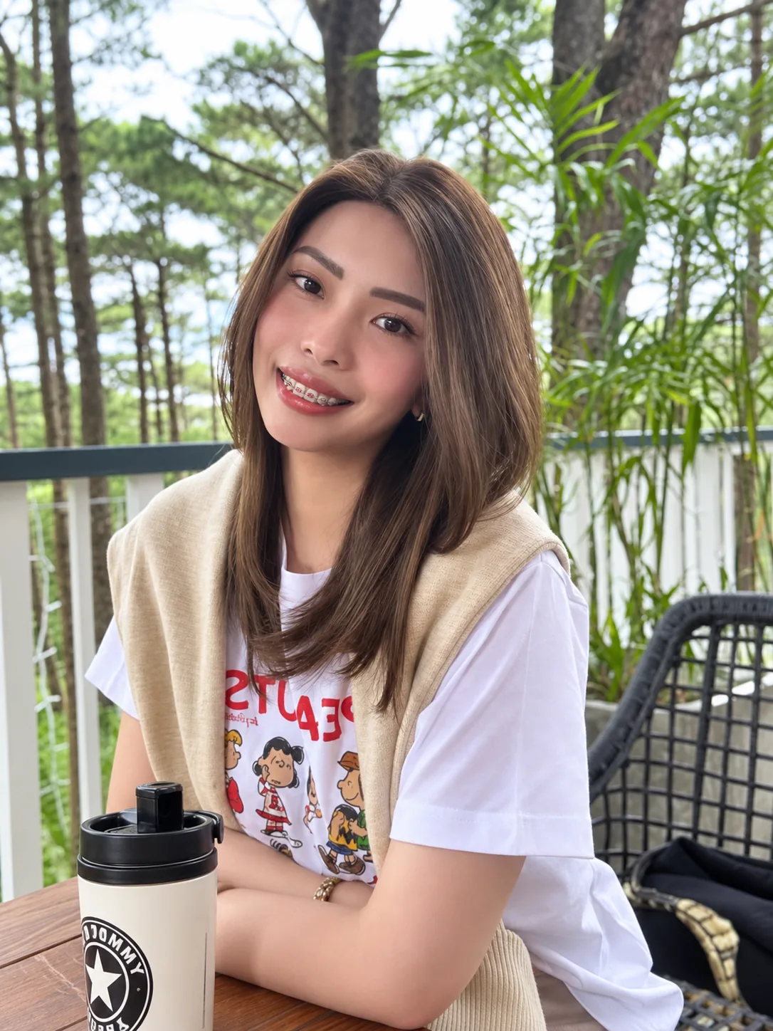 Smiling client in a brown shoulder-length human-hair wig, outdoors among pine trees